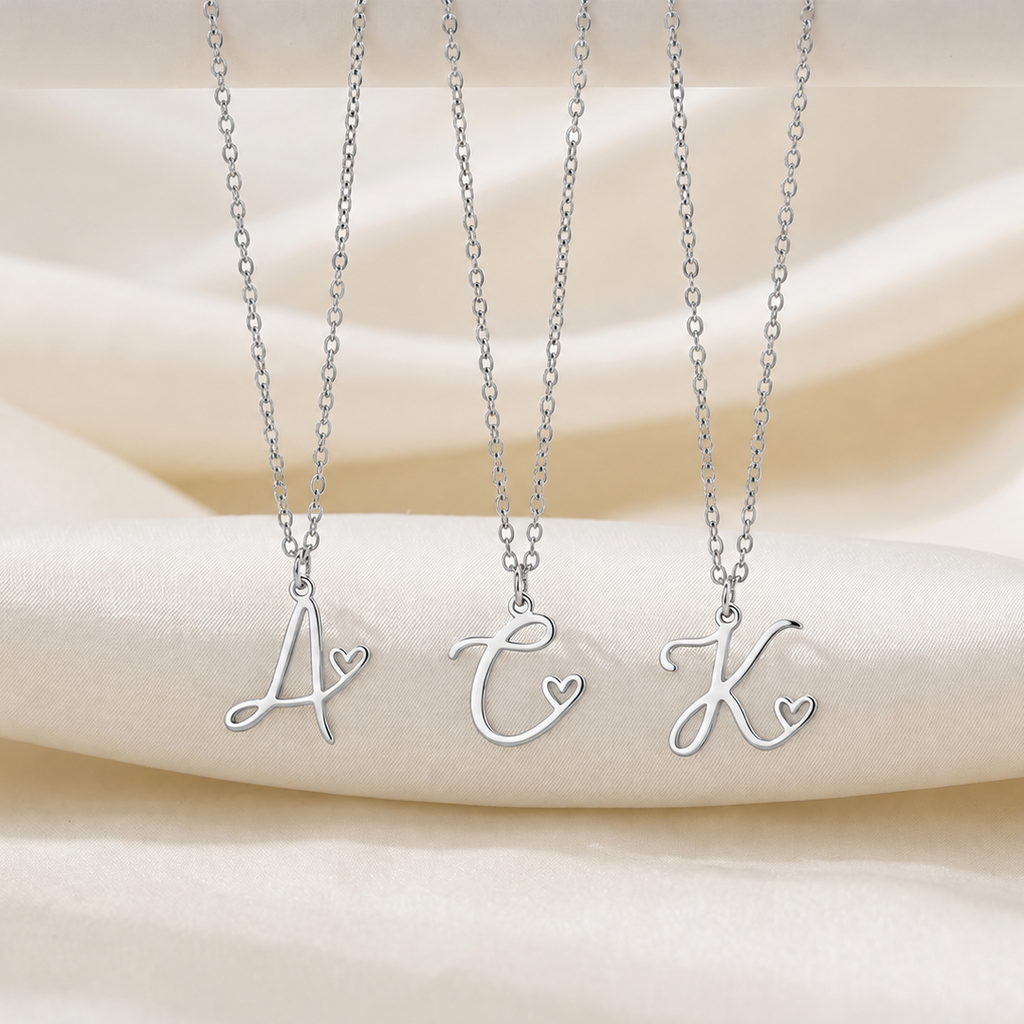 Three silver initial necklaces with cursive letter pendants (A, C, K) each featuring a small heart detail, displayed on a draped cream satin fabric background.