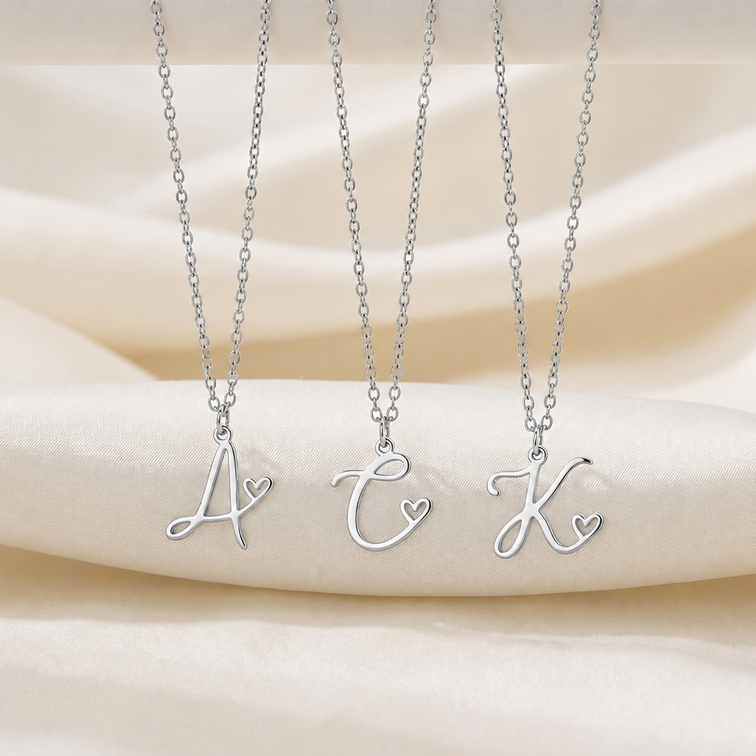 Three silver initial necklaces with cursive letter pendants (A, C, K) each featuring a small heart detail, displayed on a draped cream satin fabric background.
