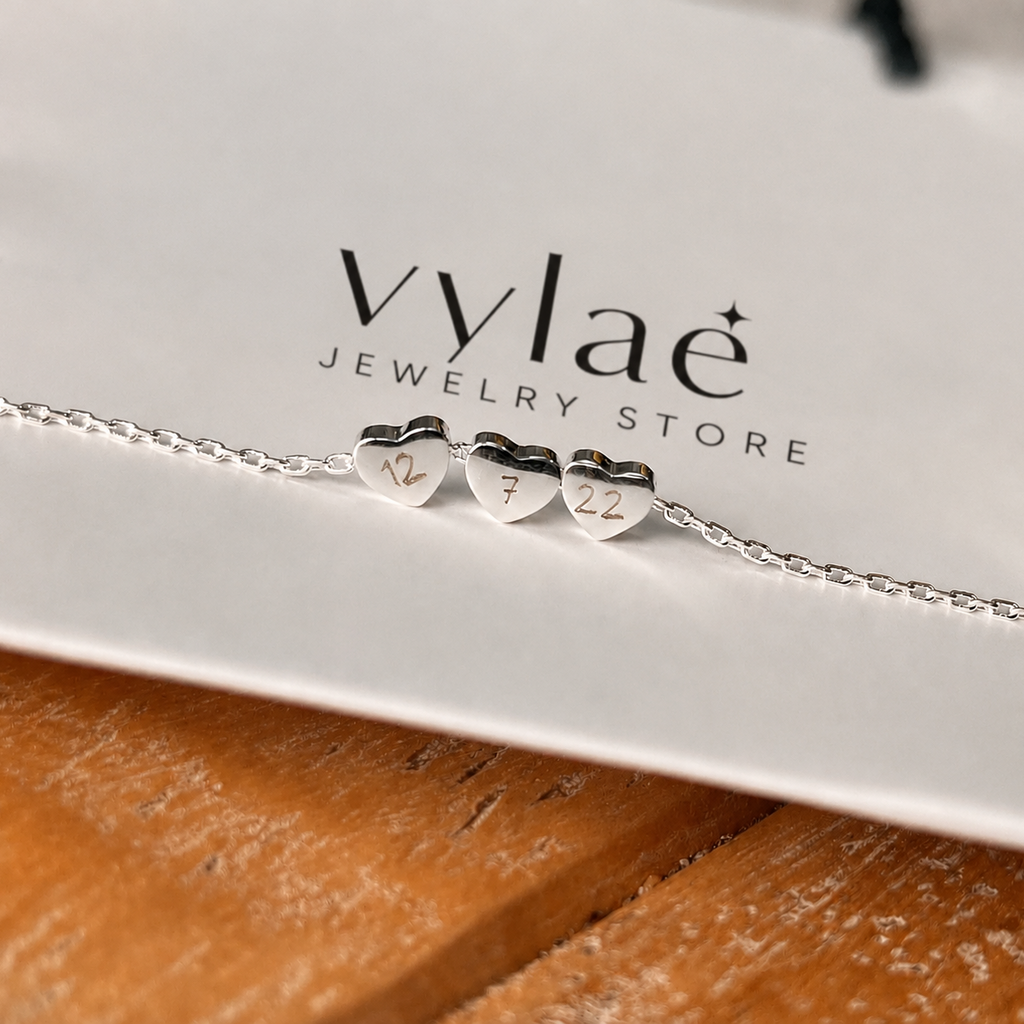 A silver chain bracelet with three small heart charms engraved with the numbers 14, 7, and 22, displayed on a white card labeled 'vylae JEWELRY STORE' over a wooden surface.