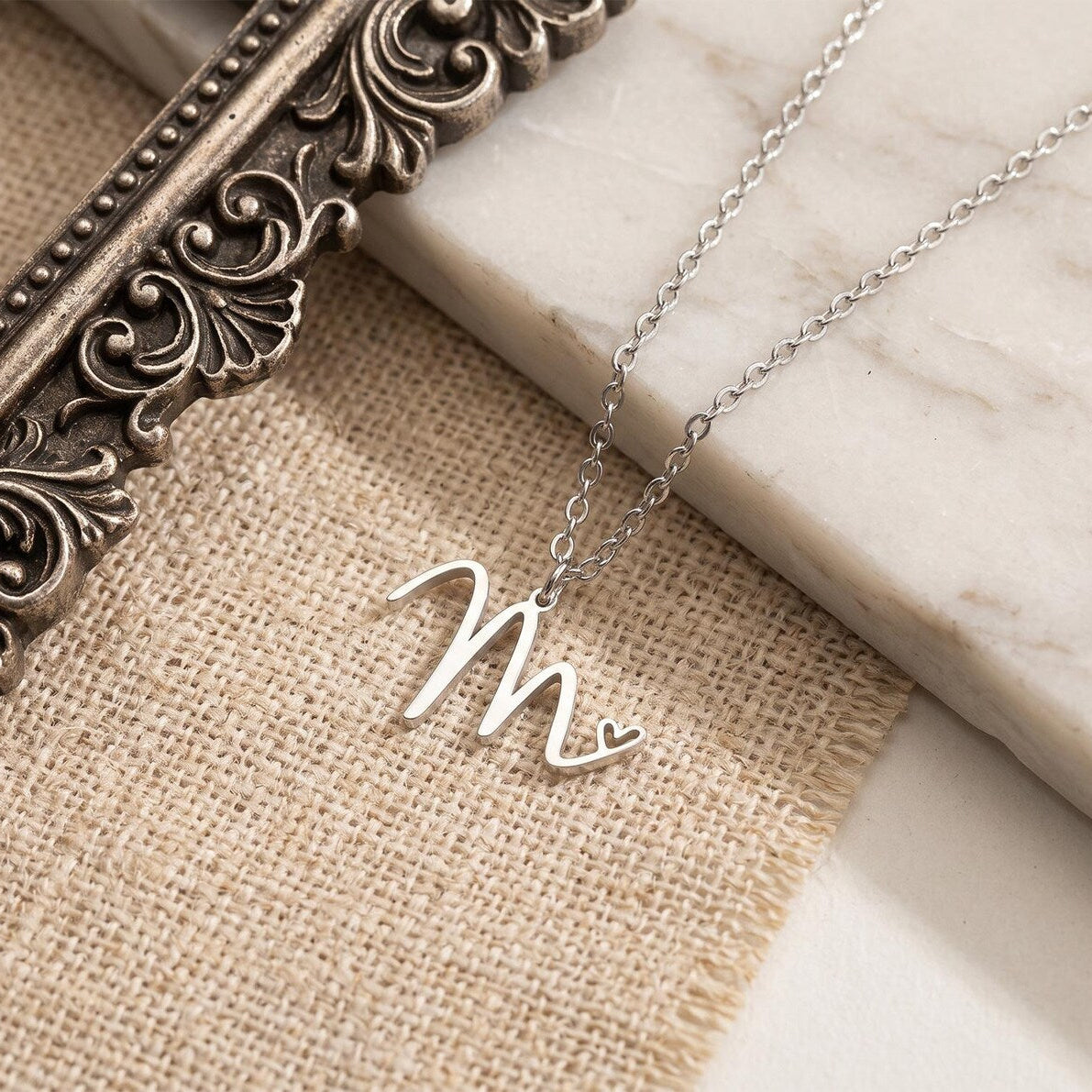 A silver script initial necklace spelling 'Mr' with a small heart at the end, displayed on a burlap fabric and white marble surface next to an ornate bronze picture frame.