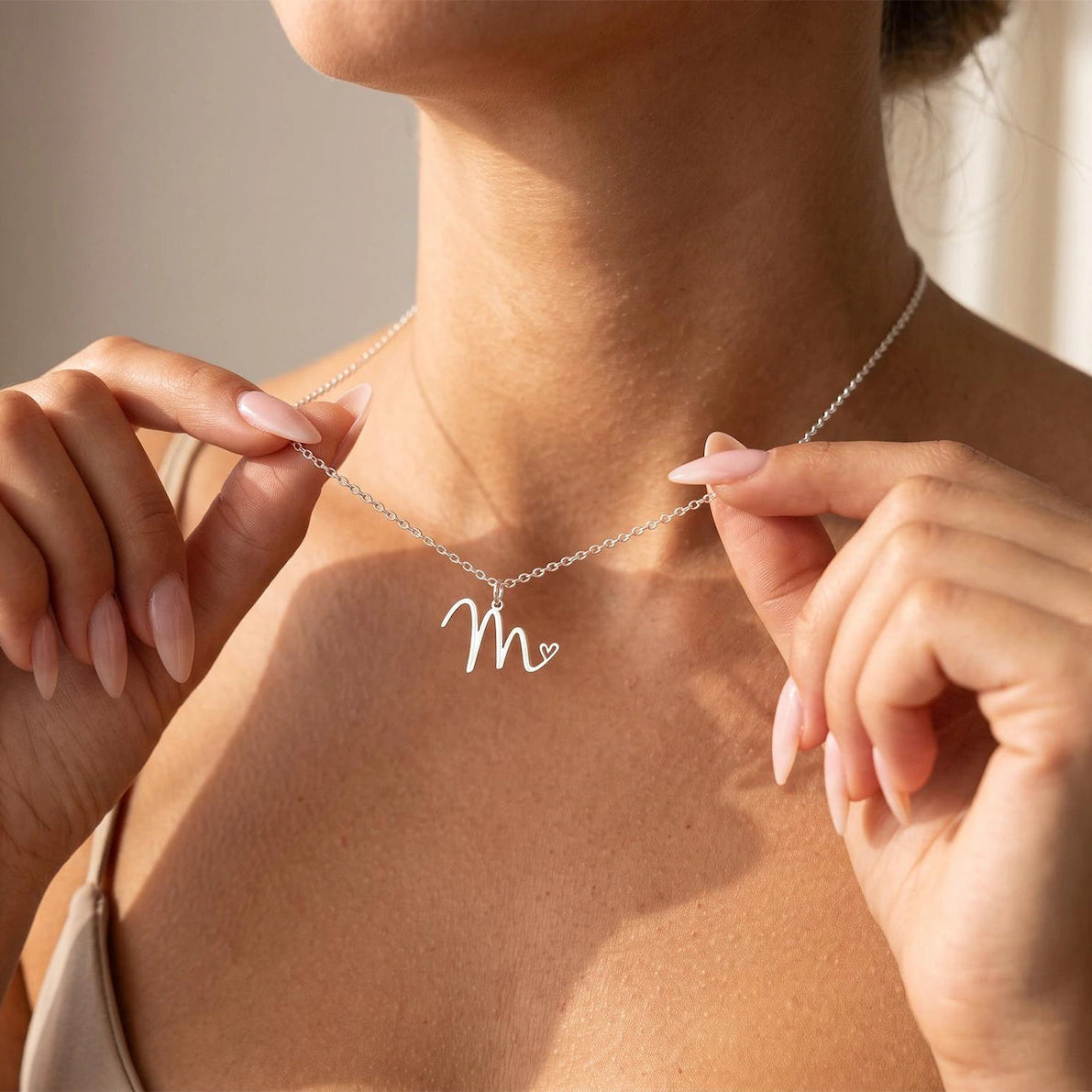 A close-up lifestyle shot of a woman wearing a delicate silver necklace with a cursive initial 'M' pendant, adjusting it with her hands.